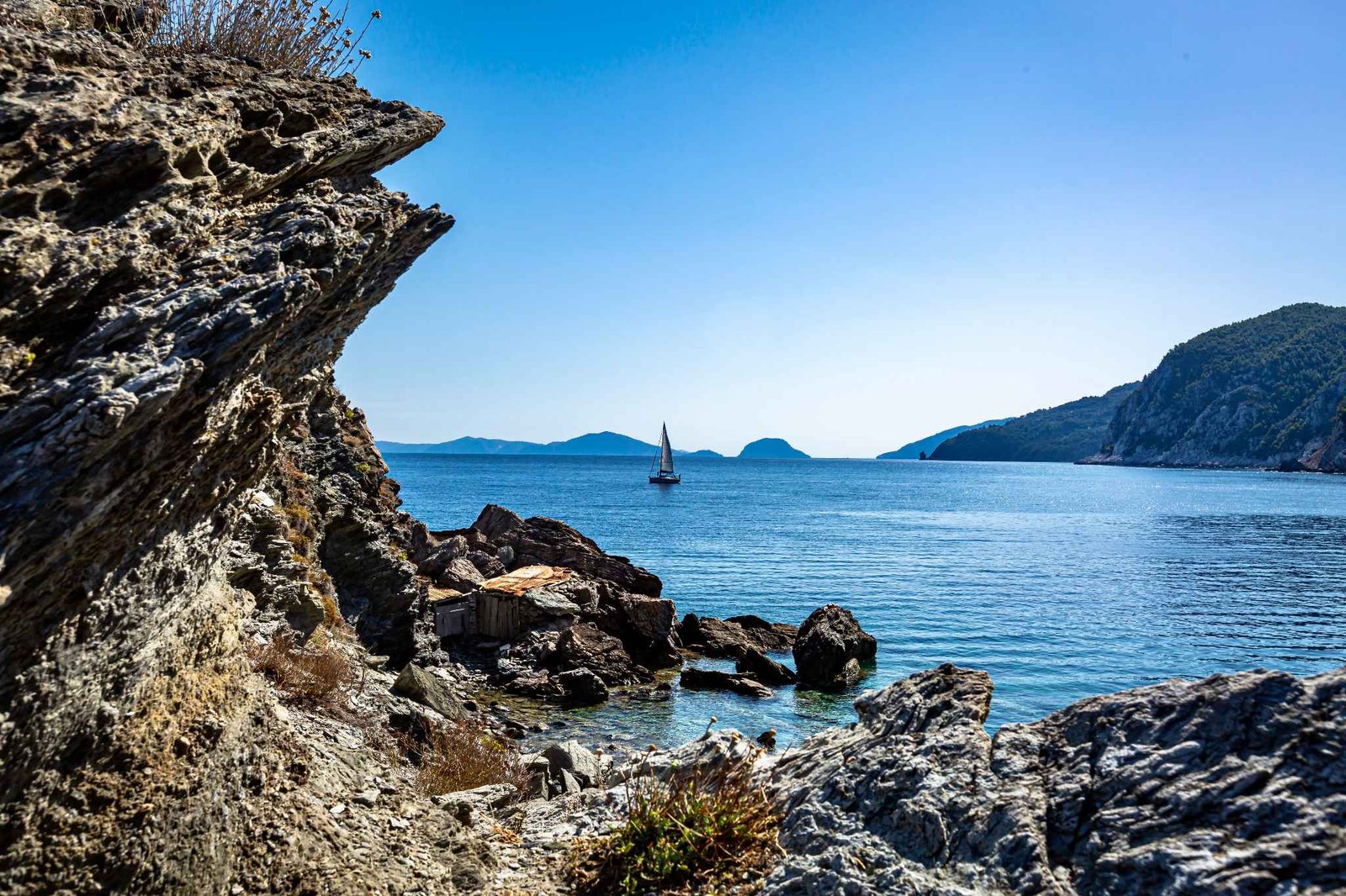 Sailboat passing a rocky cove with a wooden hut
